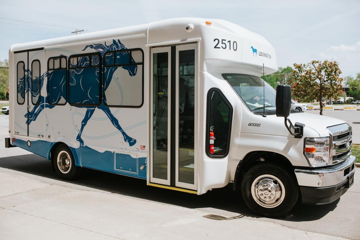 A white LexRide shuttle bus with a blue horse graphic on the side, parked in a lot on a clear day.