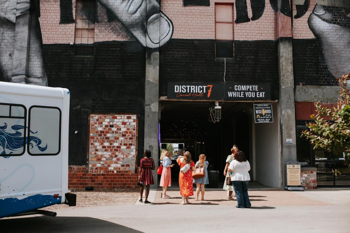 A group of people standing outside the "District 7" entrance at the Distillery District in Lexington, with a large black-and-white mural of hands visible on the building facade above them.