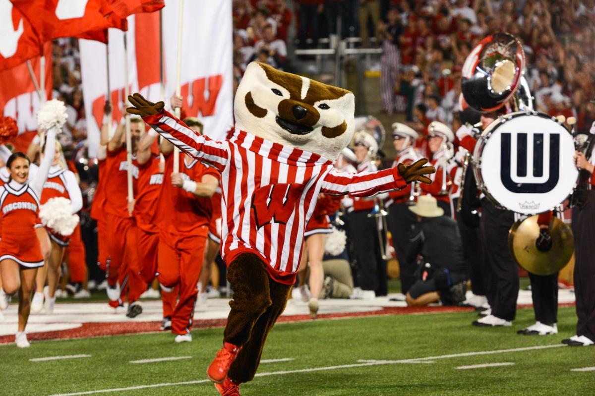 Bucky Badger runs onto the field at Camp Randall with his arms outstretched, with cheerleaders and the band behind him.