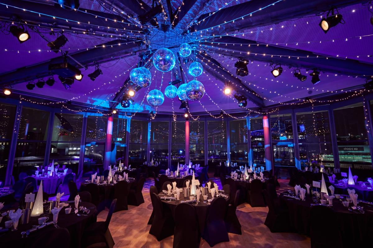 Round tables set for a festive event inside a glass-fronted venue, with blue and purple lighting overhead, hanging decorative orbs, and views of water and city lights beyond the windows.