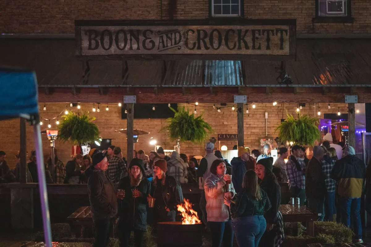 A crowd gathers at Boone & Crockett for an outdoor event at night. People in flannel and jackets socialize with drinks around a fire pit under string lights and hanging plants. The rustic bar sign hangs above the scene, creating a cozy festival atmosphere.
