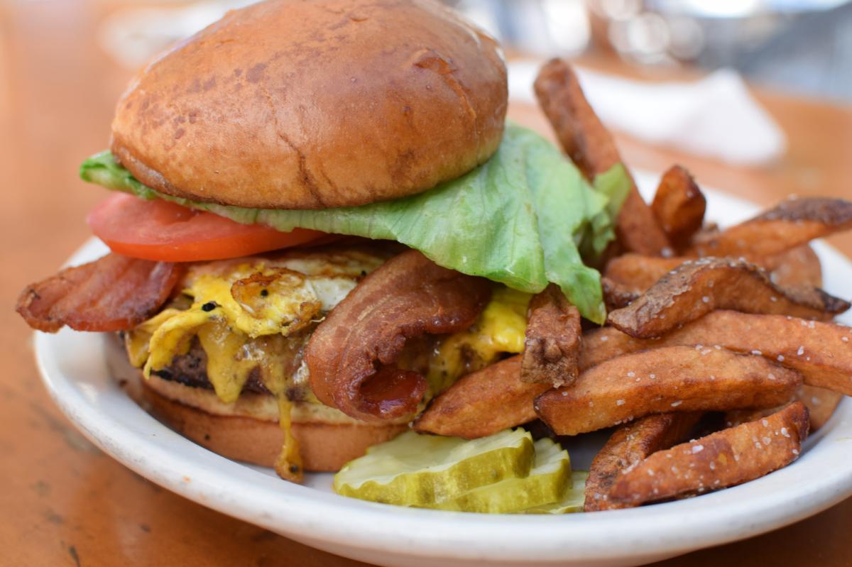 Bacon cheeseburger topped with lettuce and tomato on a brioche bun, served with seasoned potato wedges and sliced pickles on a white plate.