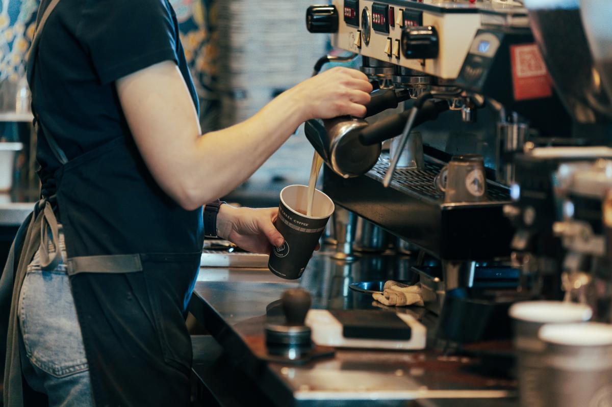 A barista pours steamed milk into a paper cup at an espresso machine inside a coffee shop.