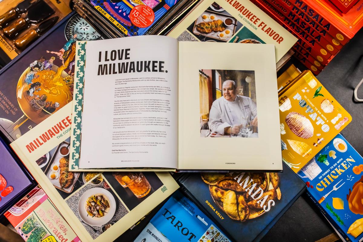 An open copy of Milwaukee Flavor: The Cookbook displayed on a table among other colorful cookbooks. The left page reads “I LOVE MILWAUKEE.” with a foreword by Chef Paul Bartolotta, and the right page shows a portrait of Bartolotta in his chef’s coat seated at a table.
