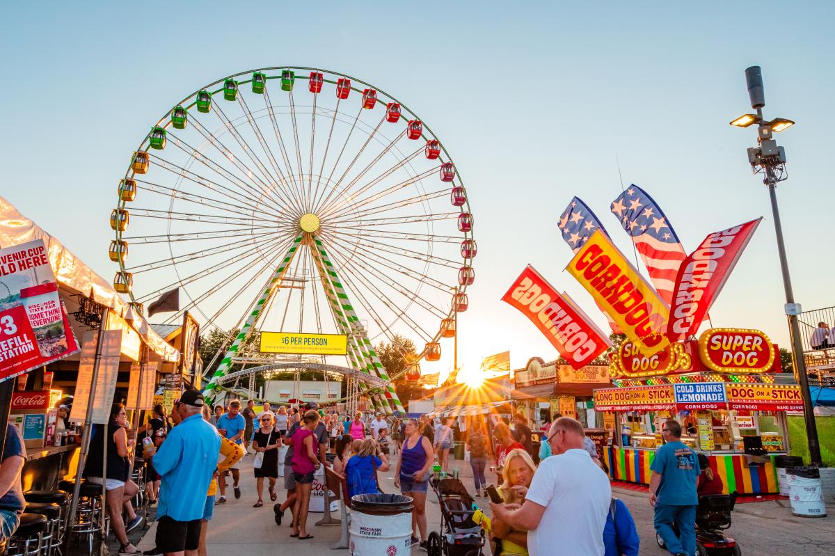 A colorful summer fair scene at sunset featuring a large Ferris wheel with green, yellow, red, and orange gondolas in the background. The foreground is bustling with people walking, eating, and enjoying the fair. Brightly lit food stands advertise corn dogs, lemonade, and "Super Dogs," with American flags waving above. A yellow sign at the Ferris wheel entrance reads "$6 PER PERSON." The sun sets behind the attractions, casting a warm glow over the festive atmosphere.