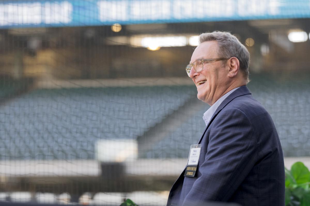 Side profile photo of a man smiling while standing at a baseball stadium. He is wearing glasses and a blue suit jacket with a name badge clipped to it. Empty stadium seats and protective netting are visible in the background.
