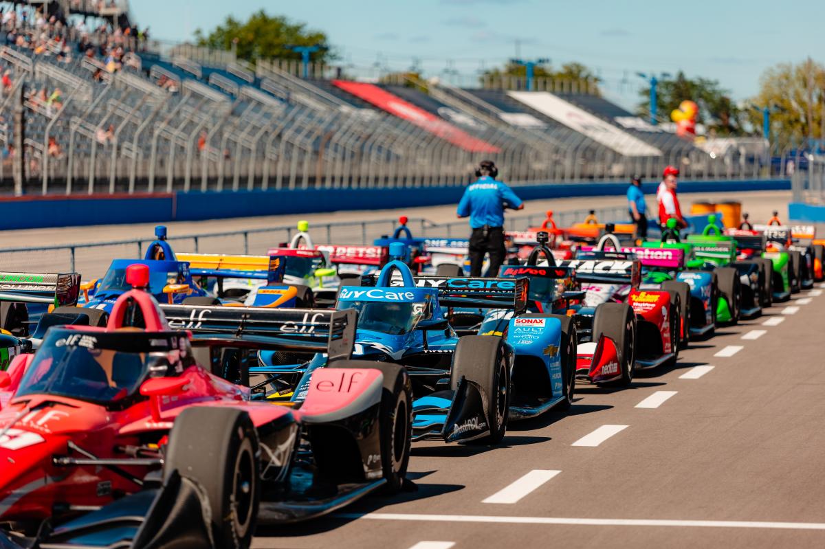 IndyCar race cars lined up on the track at the Milwaukee Mile with drivers and officials preparing for the start of the race, as spectators watch from the grandstands under a sunny sky.