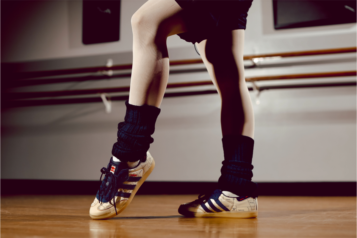 Close-up of a dancer’s legs and feet in sneakers and leg warmers, poised on a wooden studio floor near a ballet barre.