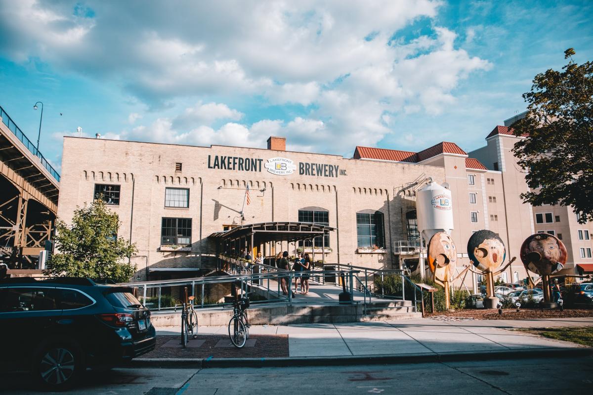 Exterior view of Lakefront Brewery in Milwaukee, a historic brick building with large windows, outdoor entrance ramp, and people gathered at the front. A white brewing tank and whimsical oversized face sculptures are visible on the right, with cars and bicycles in the foreground under a partly cloudy sky.