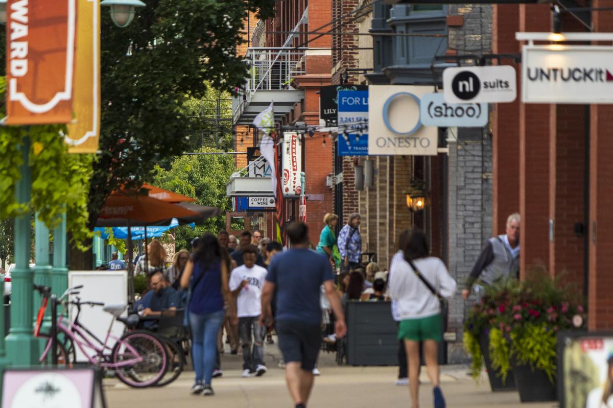 Pedestrians stroll past shops and outdoor dining areas along a bustling Historic Third Ward sidewalk, with teal streetlamps, storefront signs and string lights overhead.