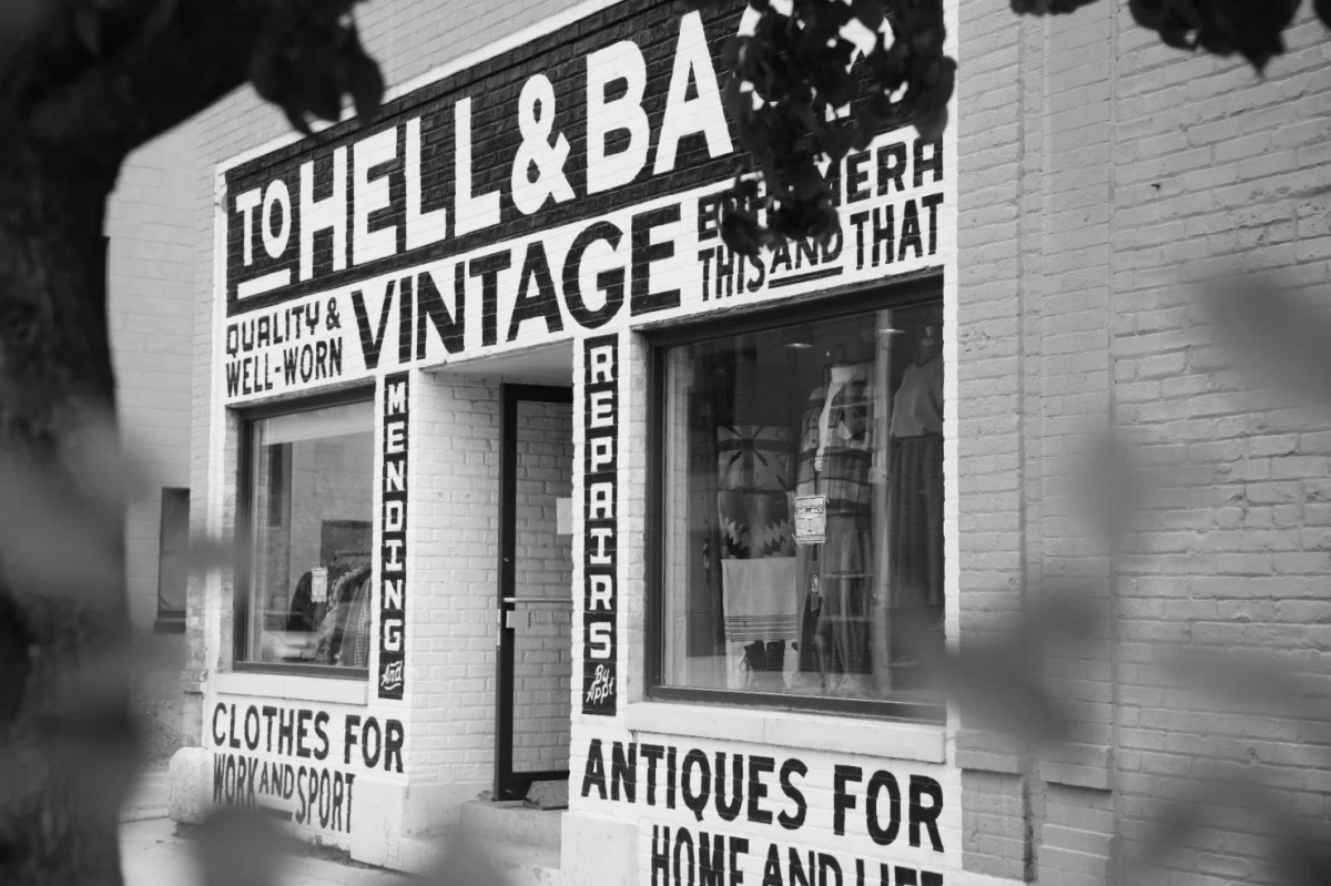 The exterior of To Hell & Back Vintage features a painted brick storefront with bold black-and-white signage advertising vintage clothing, antiques and well-worn goods. Large display windows flank the entrance, while tree branches frame the storefront in the foreground.
