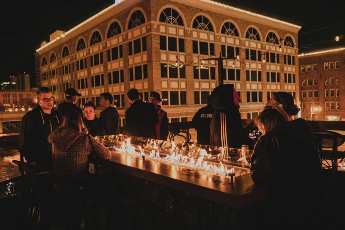 People gathered around an outdoor rooftop fire pit at night at Uncle Buck’s on 3rd in downtown Milwaukee, with a historic building lit up in the background.