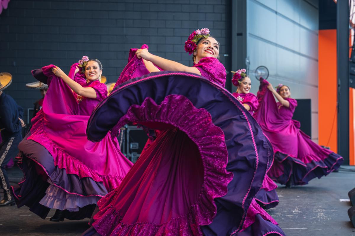 A group of female dancers performs a traditional Mexican folklórico dance on stage. They wear vibrant magenta dresses with purple ruffles and floral headpieces. The lead dancer in the foreground smiles brightly while gracefully twirling her flowing skirt. The dancers behind her mirror the movement, creating a colorful, energetic scene against a modern stage backdrop.