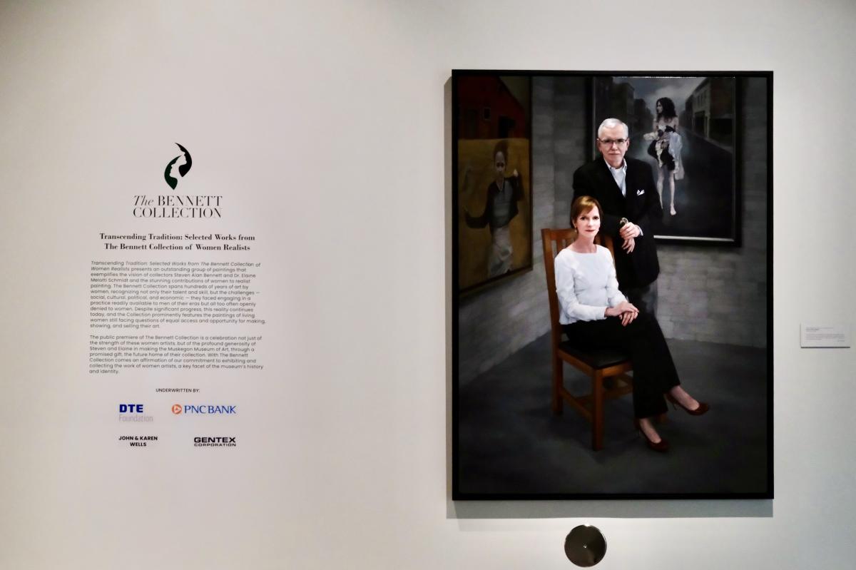Painting displayed at Bennett Schmidt Pavilion at Muskegon Museum of Art, showing Steven Alan Bennett and Dr. Elaine Melotti Schmidt, one seated and one standing, in a formal pose against a dark backdrop. Information about the collection is presented on an adjacent wall.