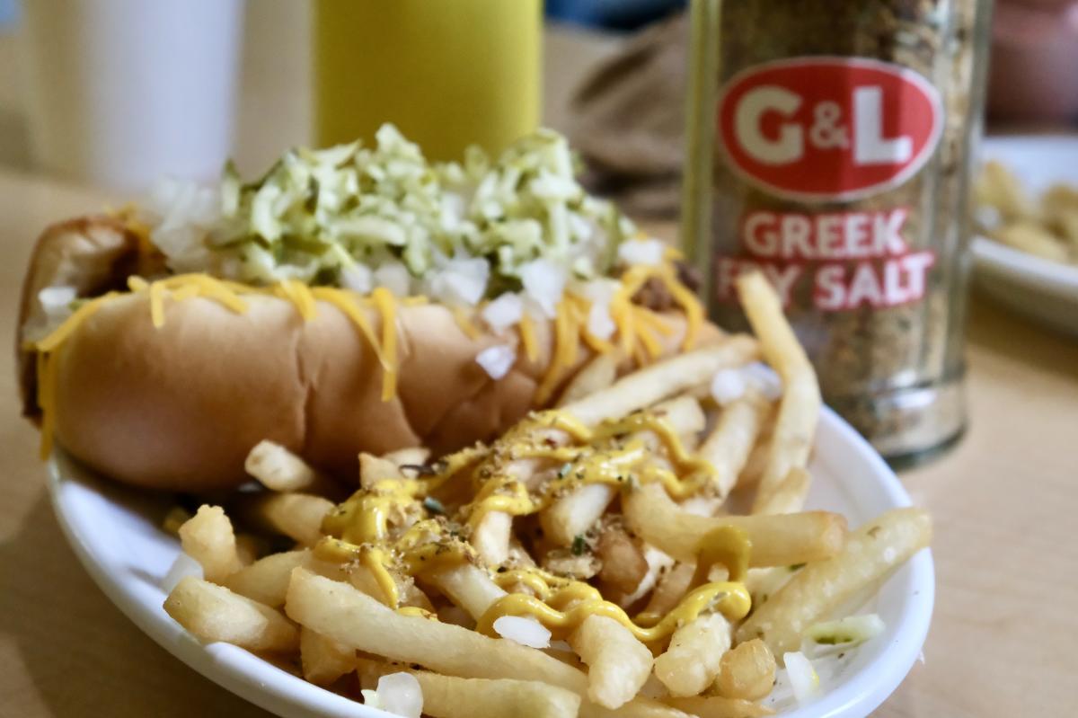 Close-up of a meal featuring a hot dog topped with mustard and onions, a side of french fries, and a container of Greek Fry Salt, all set on a table.