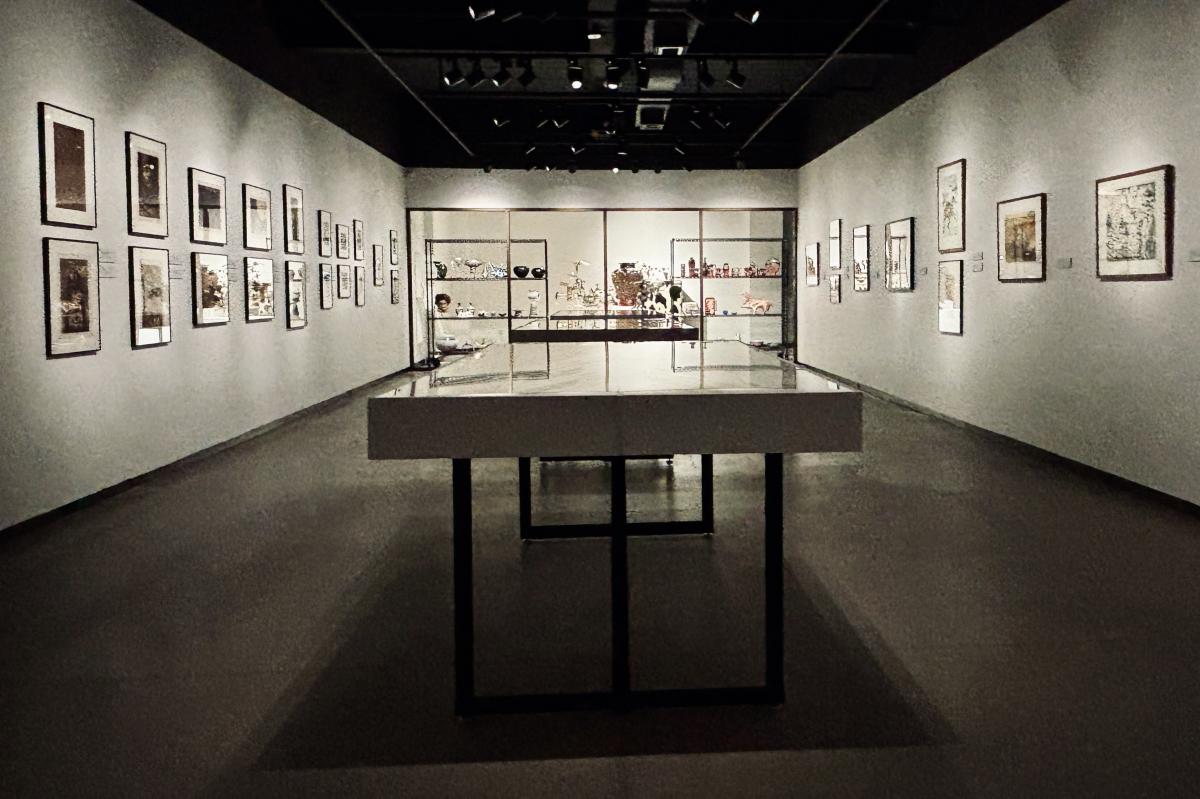 Interior of a dimly lit art gallery featuring multiple framed artworks on the walls and glass display cases with items in the center. The space is designed for an immersive viewing experience.