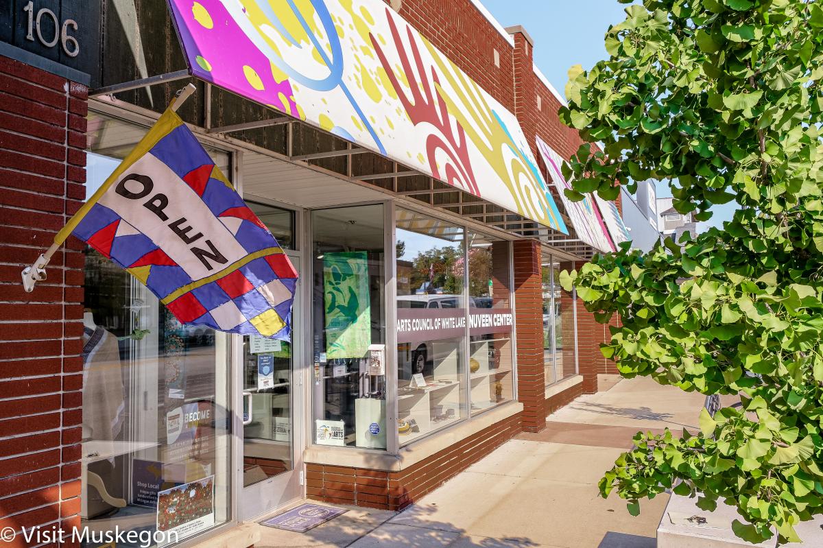 Exterior view of the Nuveen Center Arts Council of White Lake with a colorful banner overhead and an "OPEN" flag hanging beside the entrance.