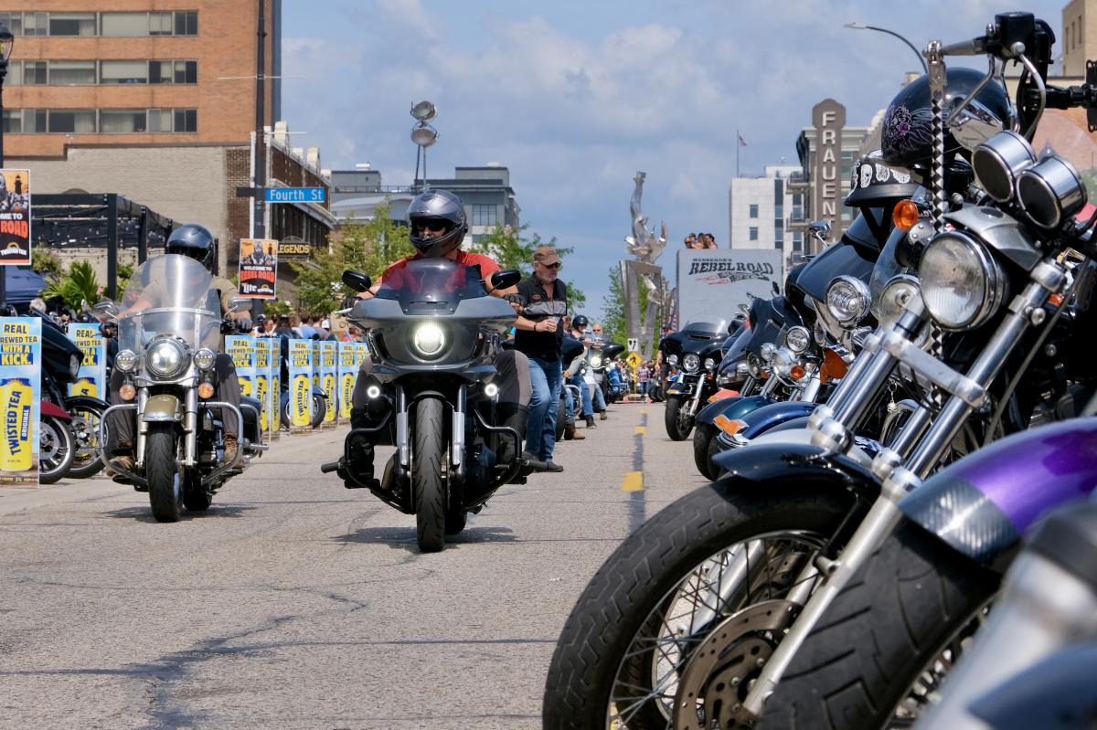 Line of parked motorcycles on a city street with a person riding a motorcycle in the background.