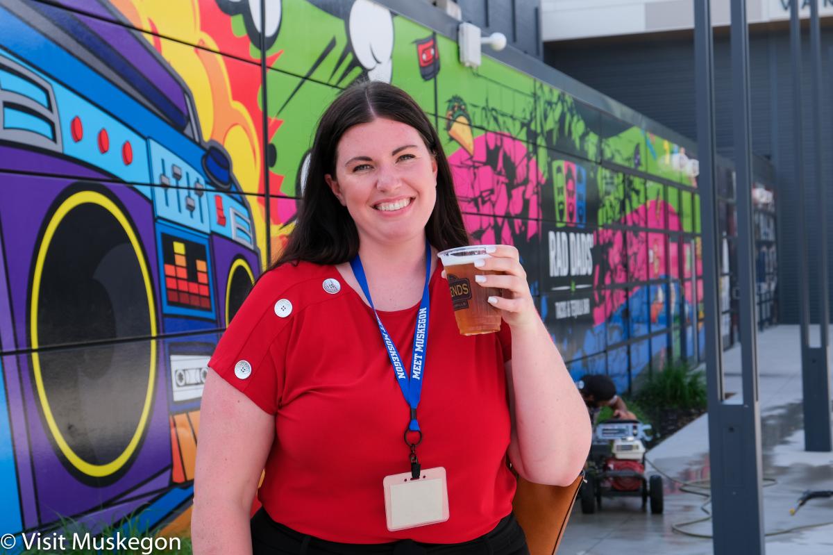 Smiling woman with long dark hair in red shirt wears blue lanyard. She holds social district cup of beer up and stands in front of mural