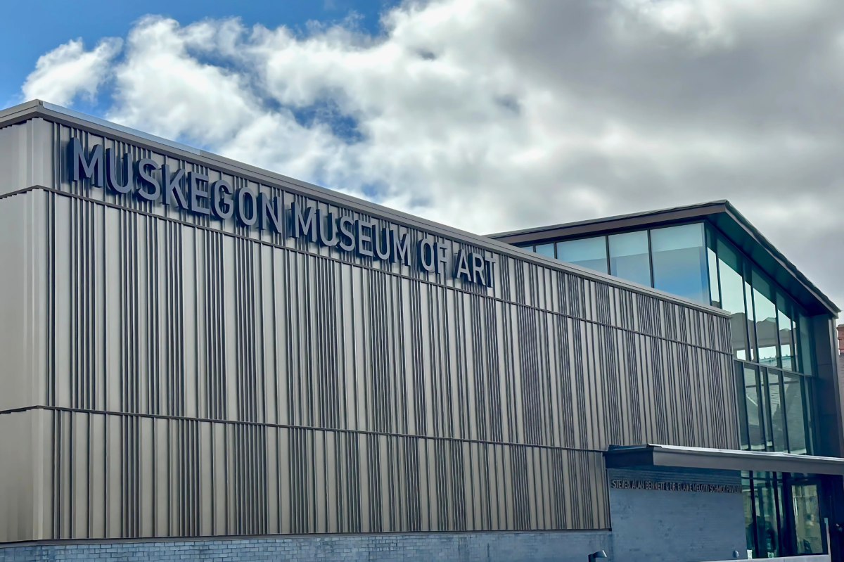 Exterior view of the Muskegon Museum of Art under a partly cloudy sky. The building features a modern design with elongated vertical panels and large glass windows.