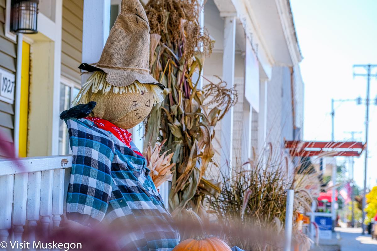 scarecrow in blue plaid shirt sits among fall decor in front of business