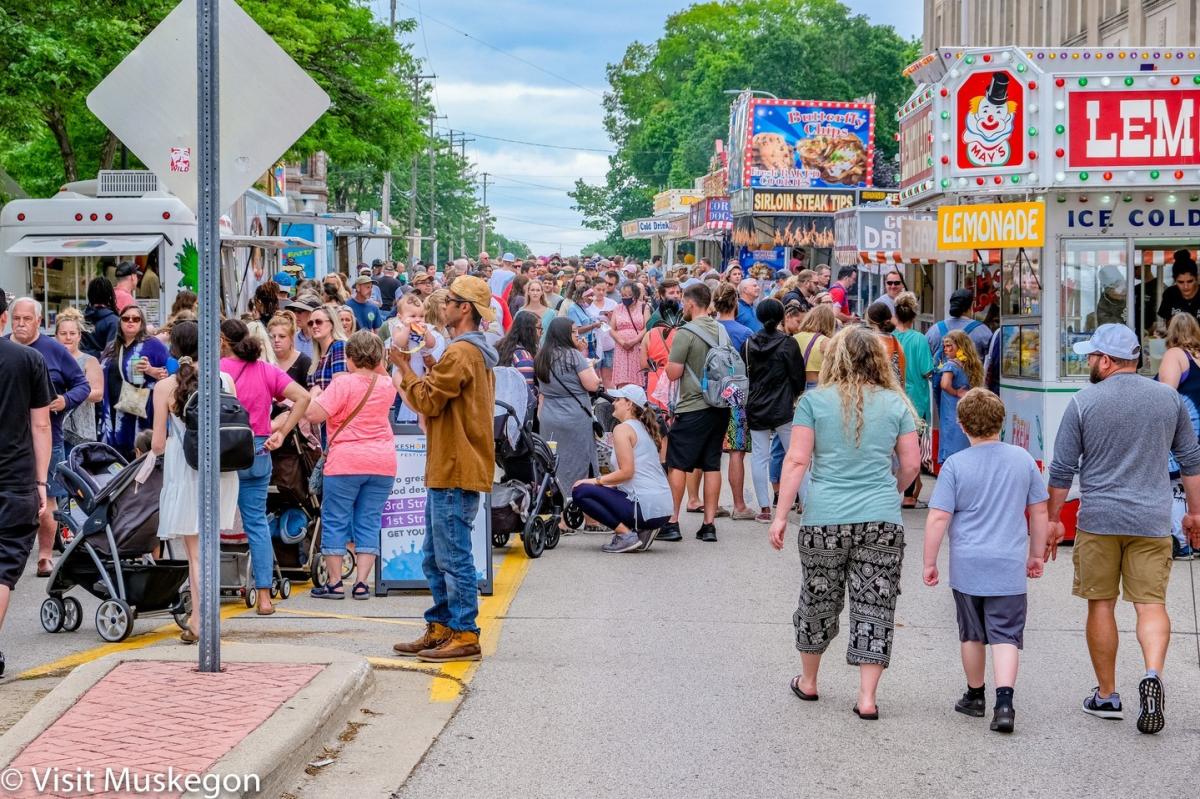 A bustling street scene at a festival in Muskegon with a crowd of people walking, talking, and visiting food stands offering snacks like lemonade and popcorn under a cloudy sky.