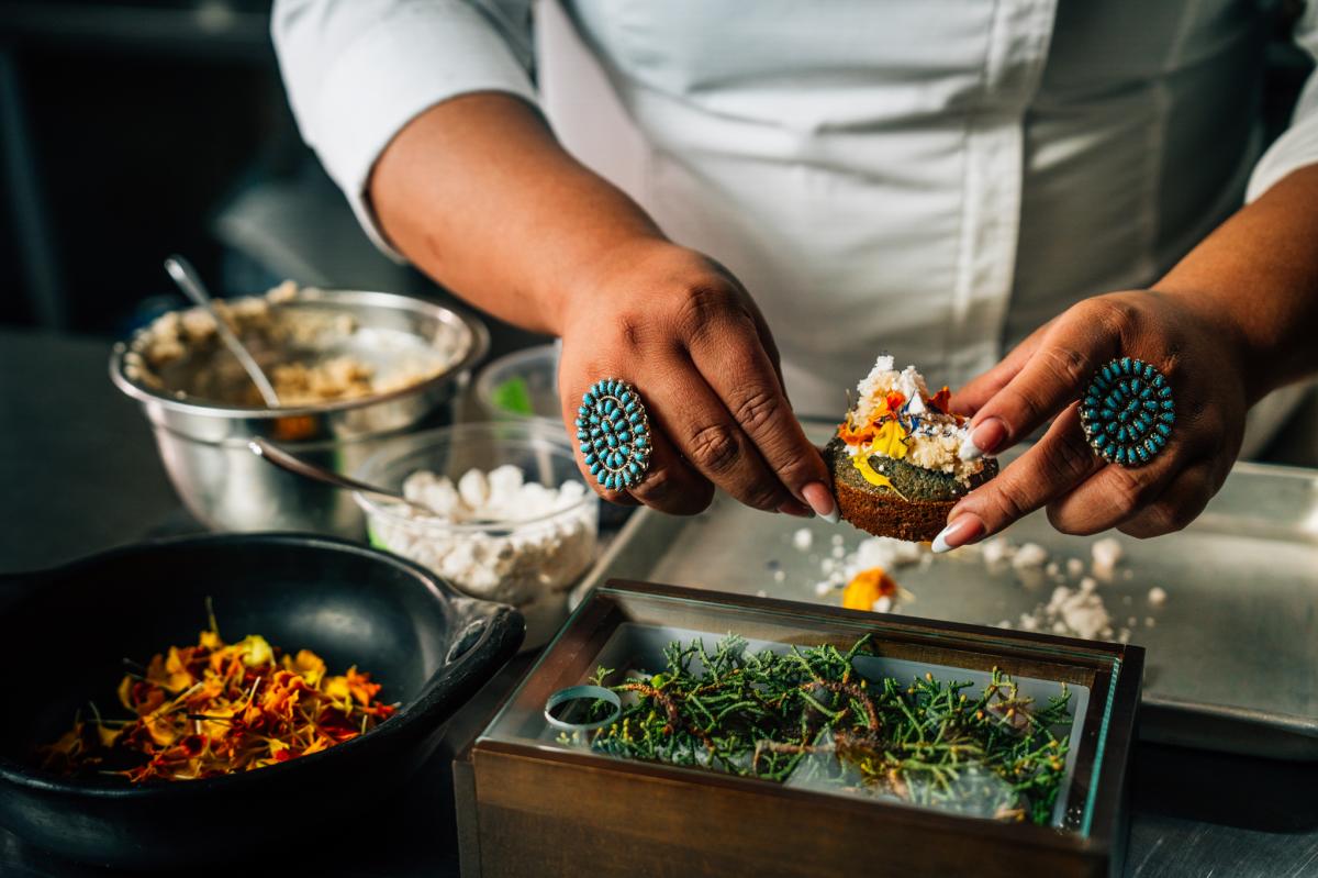 Person preparing gourmet food in a professional kitchen, adding toppings to an open-faced sandwich, surrounded by various fresh ingredients.