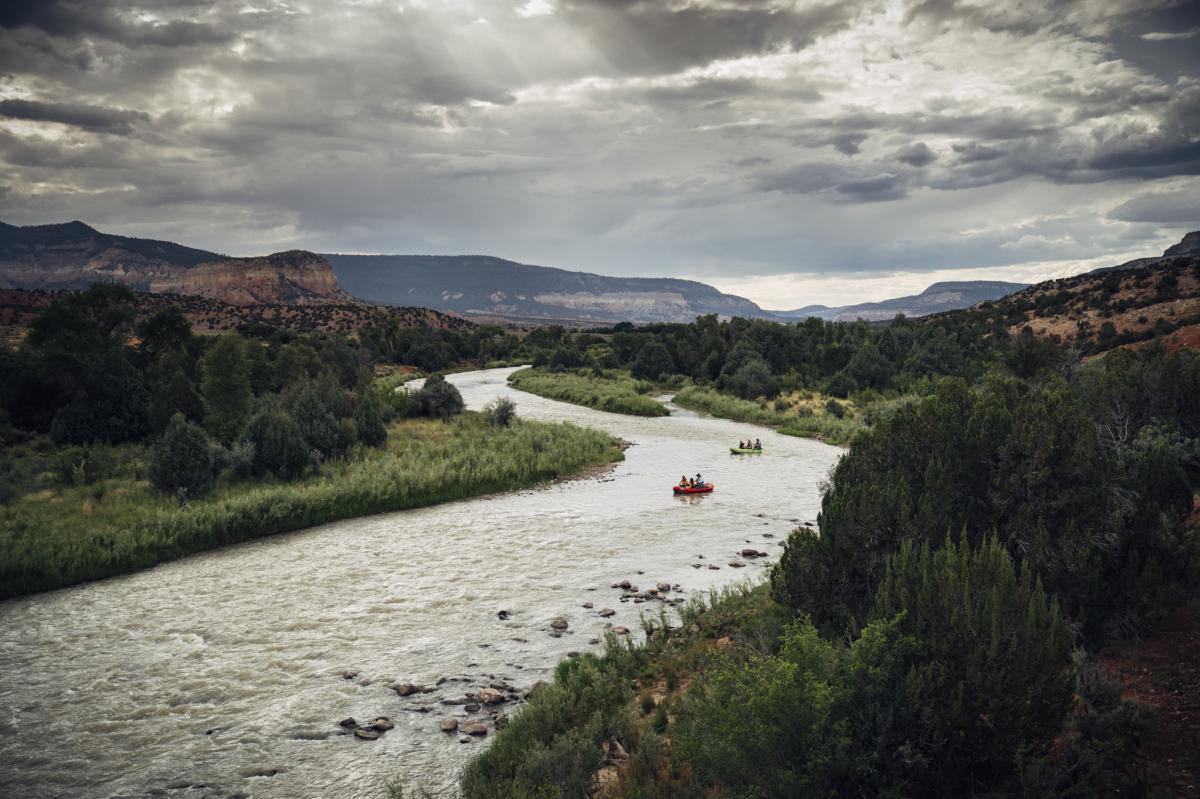Rafters navigate a winding river through a scenic desert landscape under a cloudy sky.