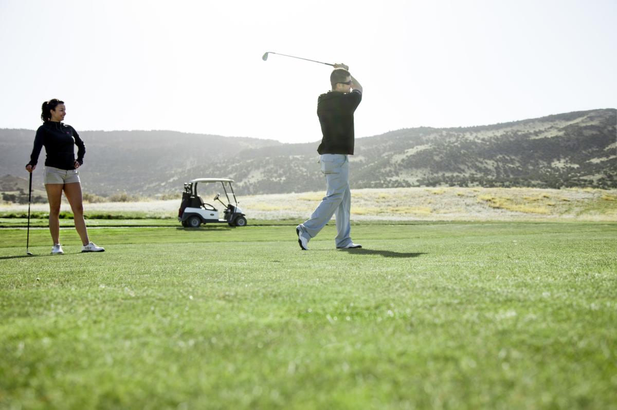 A person watches another person who is mid swing on a green golf course surrounded by hills with a golf cart in the background