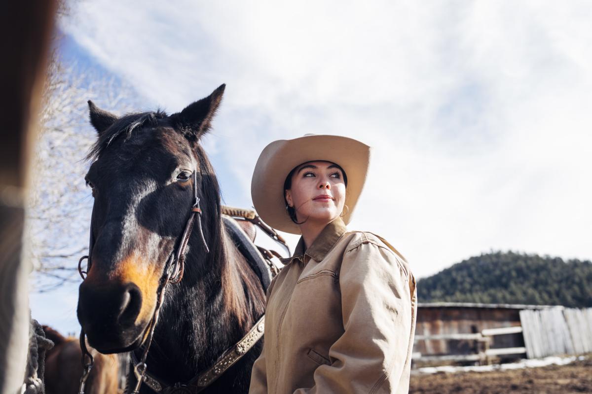 A person wearing a cowboy hat stands beside a dark brown horse. The sky is clear with a few clouds, and there is a wooden fence and tree in the background.
