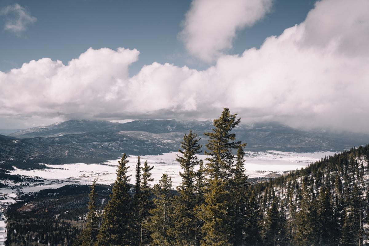 View of a snow-covered landscape with tall pine trees in the foreground and mountain ranges under a cloudy sky in the background.