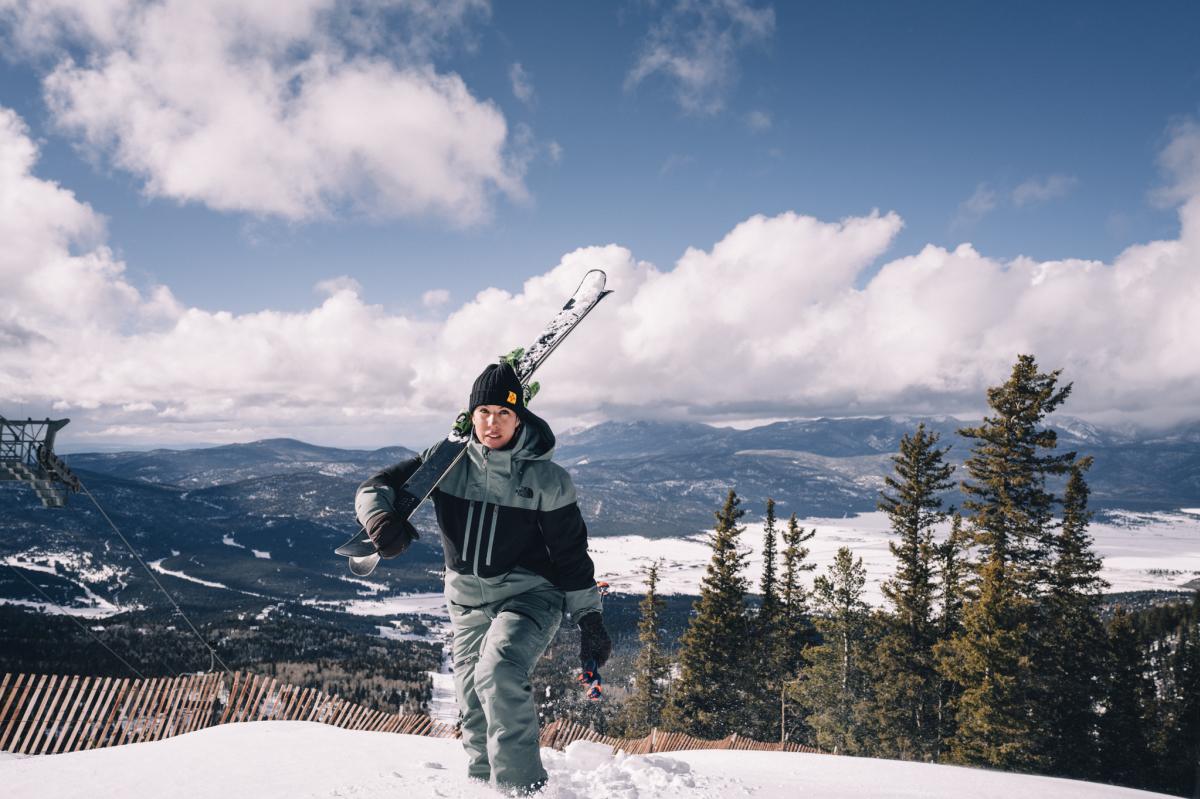 Person carrying skis over their shoulder walking in the snow with a scenic mountain landscape in the background.