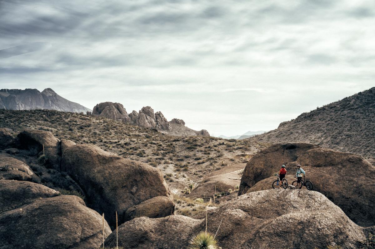 Two people with bicycles standing on a rocky terrain in a desert landscape, with mountains in the background under a cloudy sky.
