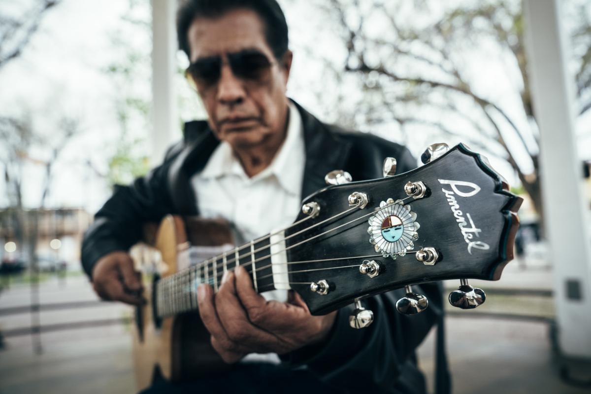 Person wearing sunglasses playing a Pimentel acoustic guitar outdoors.
