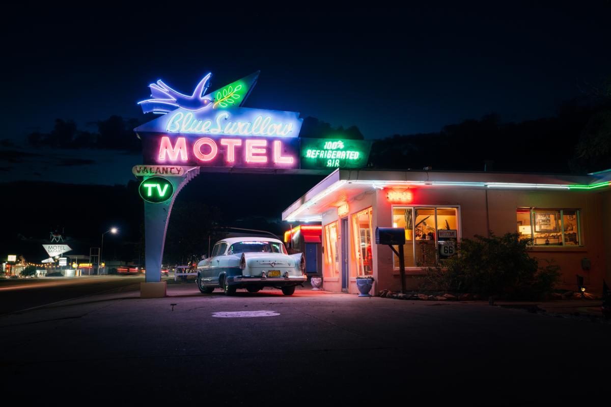 Night view of the Blue Swallow Motel with vibrant neon signs, featuring a classic car parked in front.