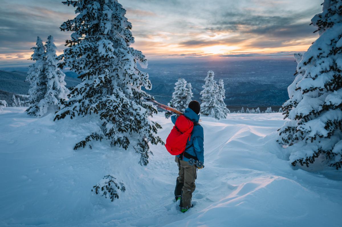 A person stands on the summit of a snowy mountain overlooking the landscape with a red backpack and skis slung over their shoulder