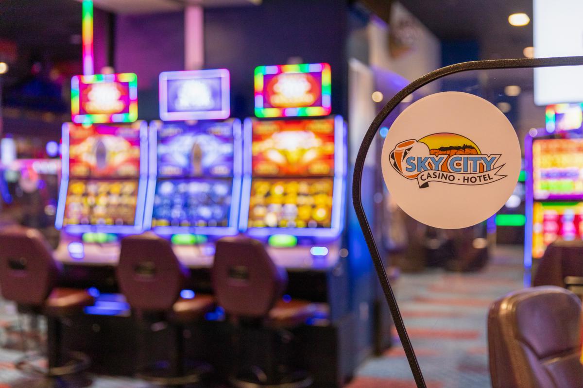 Sign with "Sky City Casino - Hotel" logo in focus, with blurred background of colorful casino slot machines and seating area.