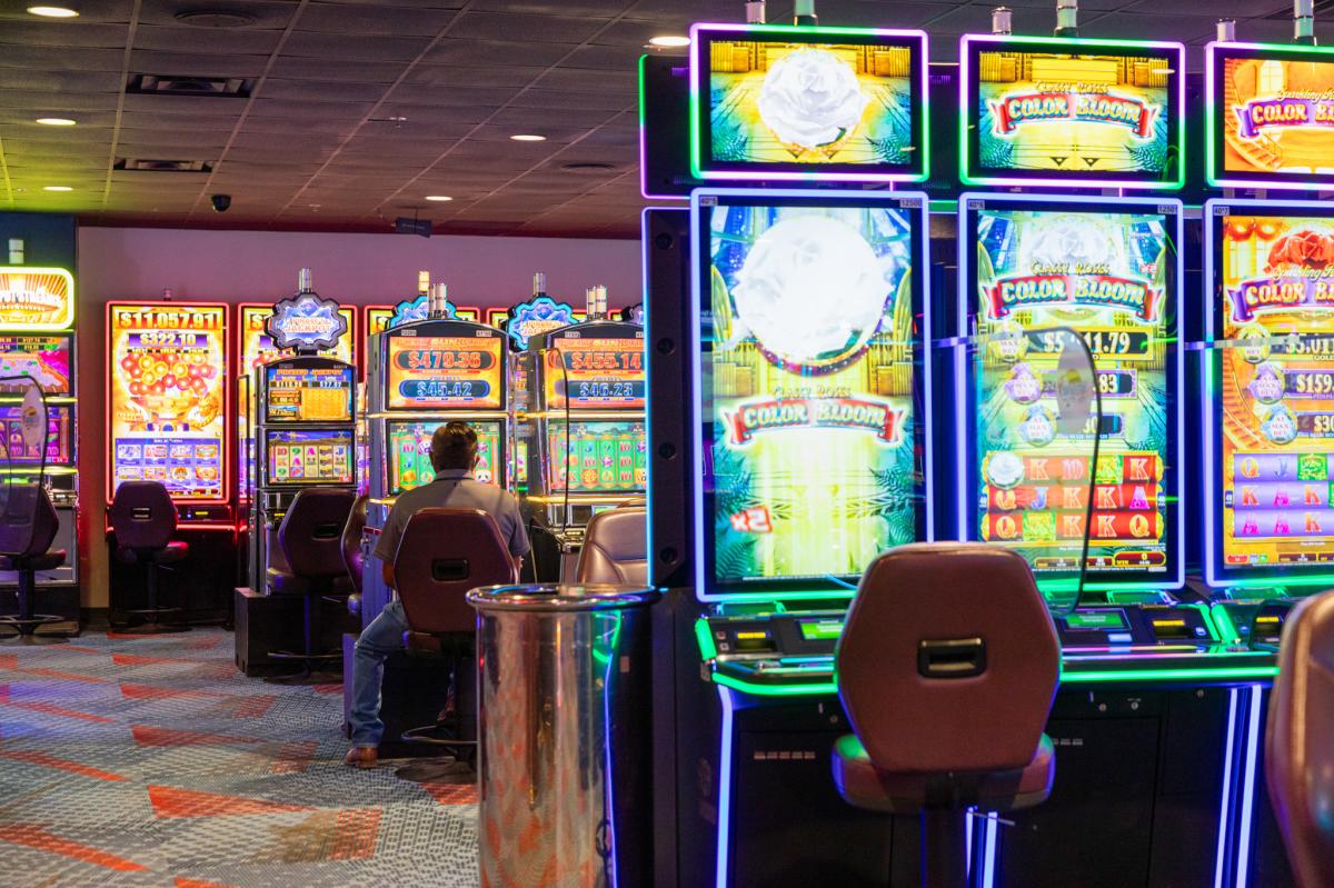 Interior of a casino showing vibrant slot machines with colorful displays. A person is seated at one of the machines in a casual setting.