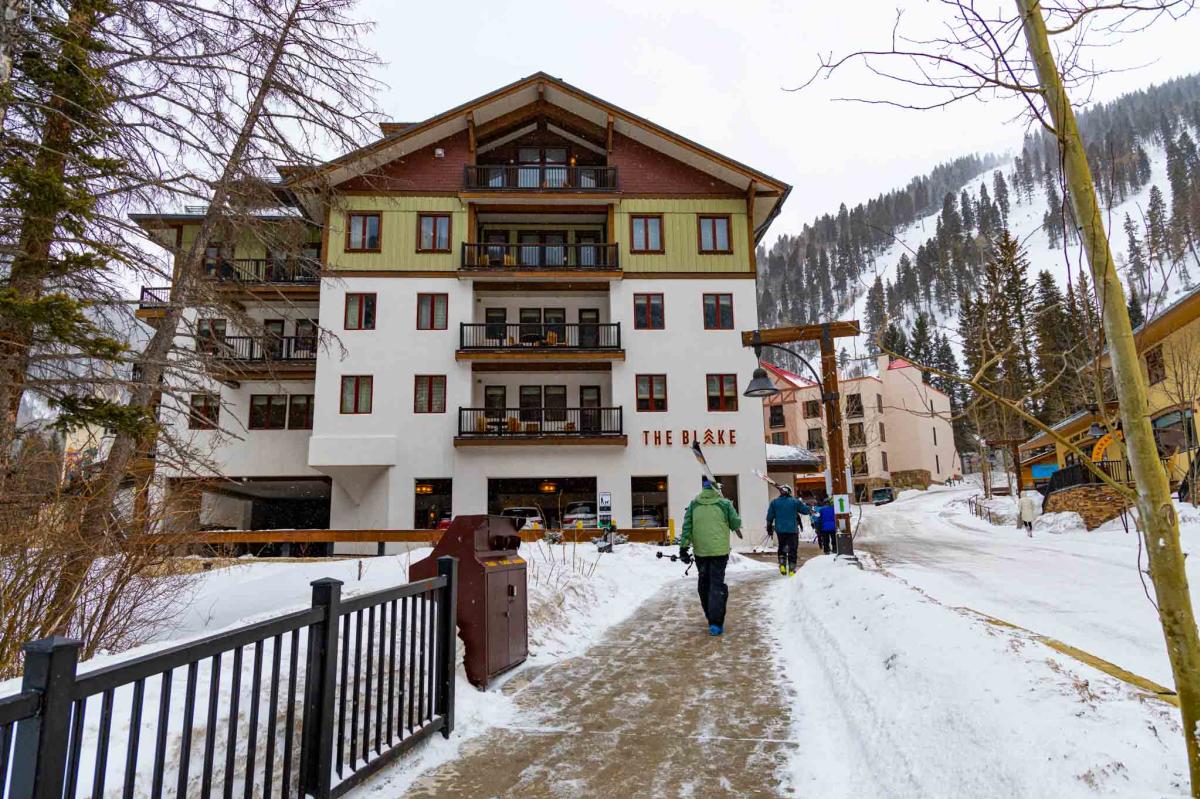 A multi-story hotel labeled "The Blake" sits next to a snow-covered mountain with people holding skis walking next to it.