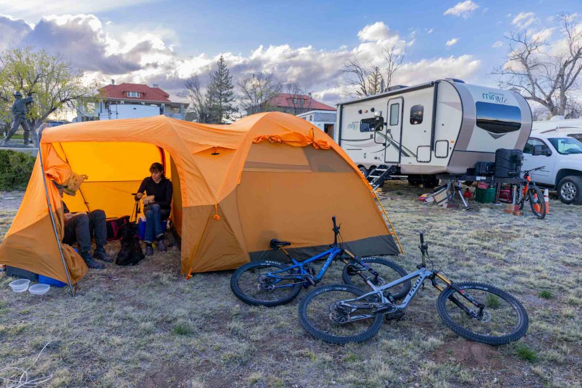 A person sitting inside an orange tent next to a small trailer and bicycles, in a camping area during dusk.