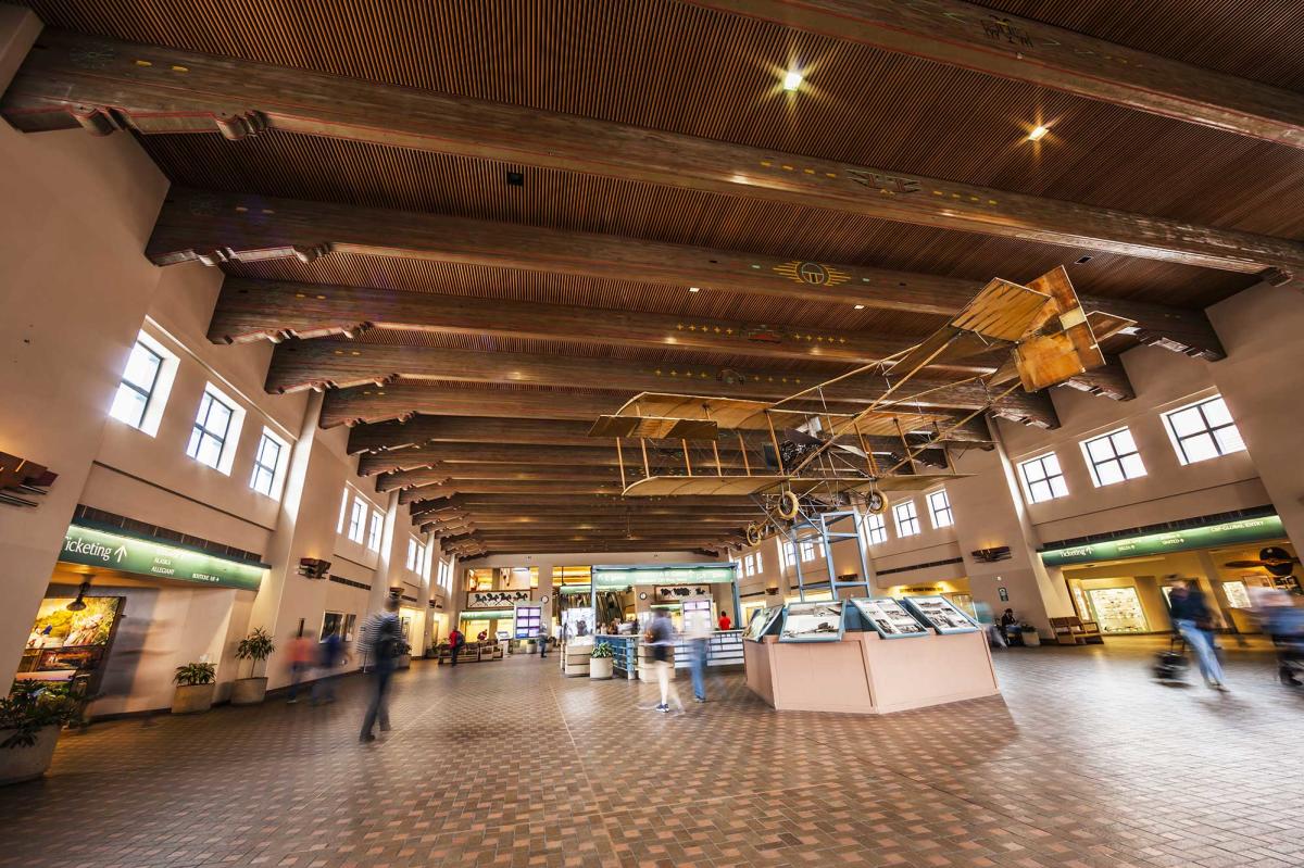 A spacious airport terminal with wooden beams and a vintage aircraft model suspended from the ceiling. The floor is tiled, and there are display cases and people walking in blurred motion.