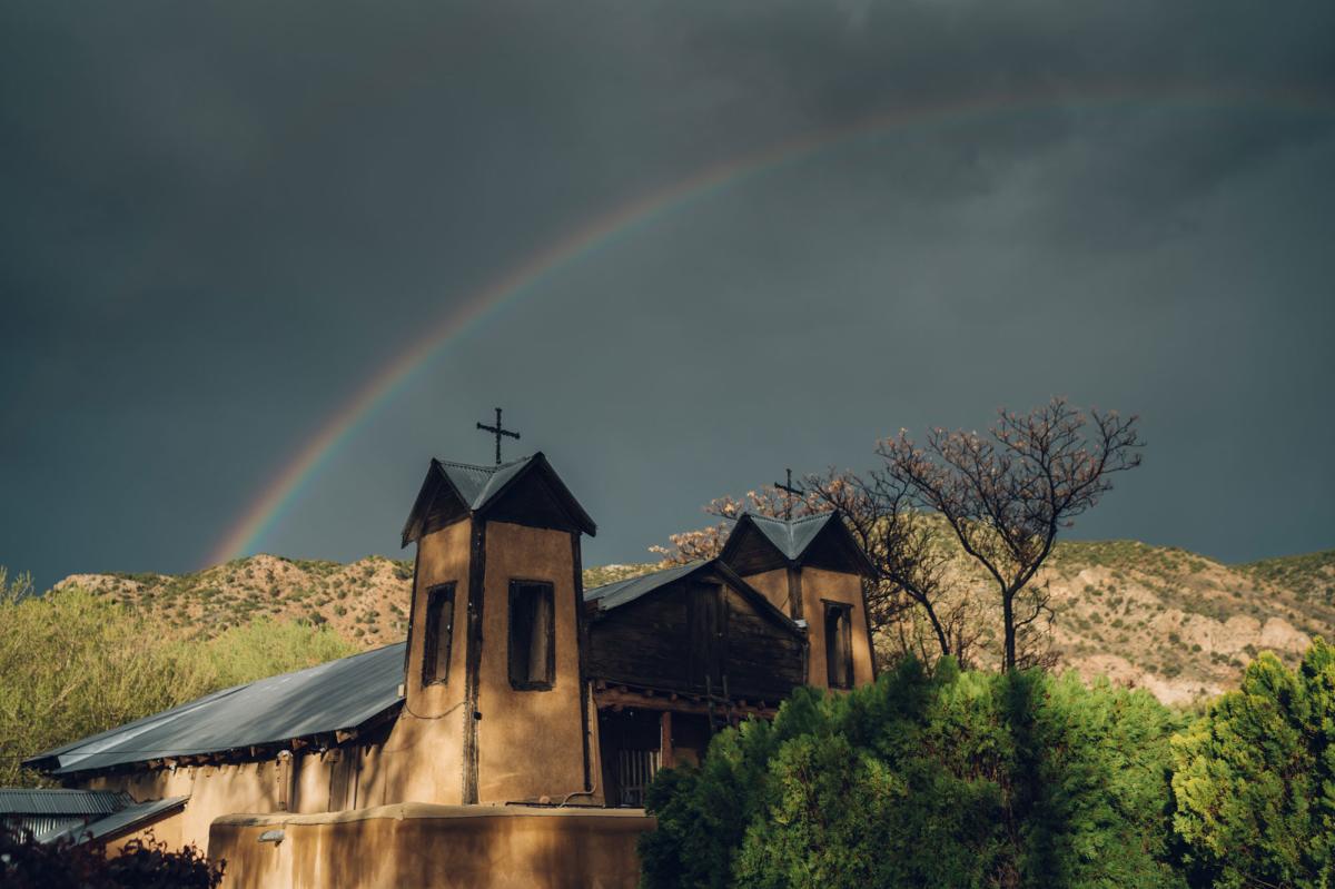 A rainbow arcs over a traditional adobe church with two steeples, both with crosses on top