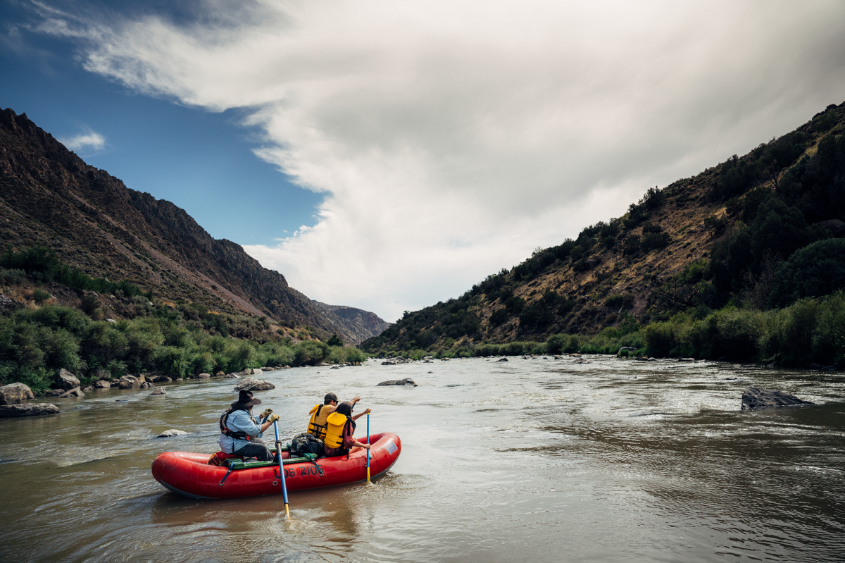 two people sit in a red raft as they paddle down a flowing river between two sprawling hills on either side