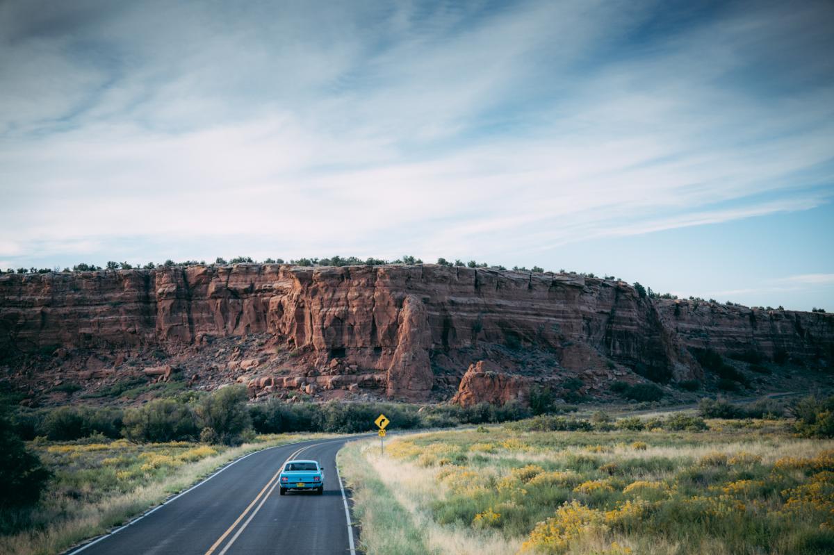 A car drives on a winding road past a large red rock formation under a clear blue sky.