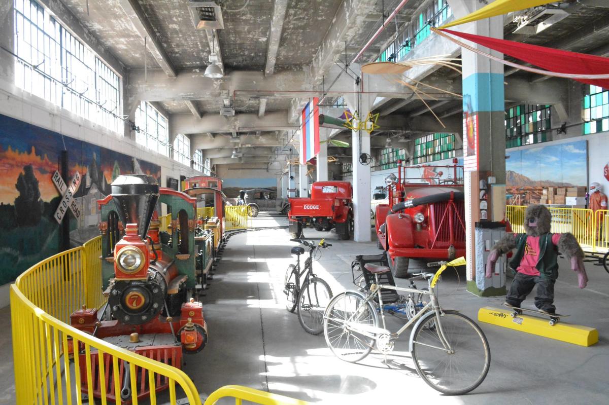 A museum exhibit features vintage vehicles, including a red and green mini steam locomotive and red trucks.
