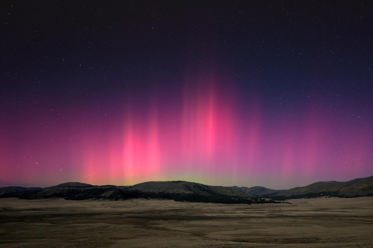 A vibrant aurora, with pink and green lights, illuminates the night sky above dark rolling hills.