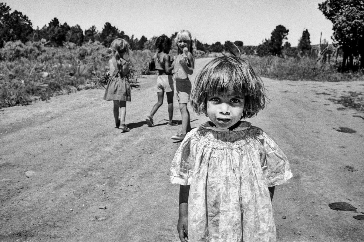 Black and white photograph of a small child standing in the foreground of a dirt road.