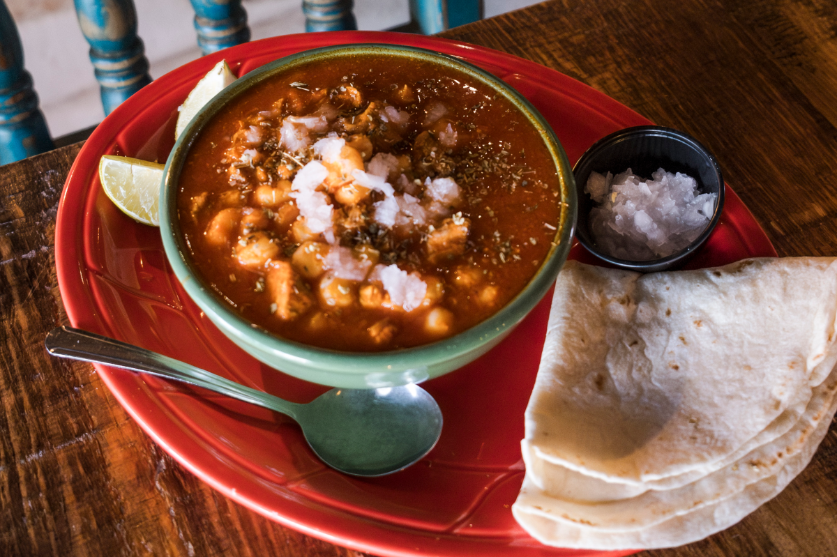 A bowl of red posole sits on a red plate with a side of corn tortillas and chopped white onion.