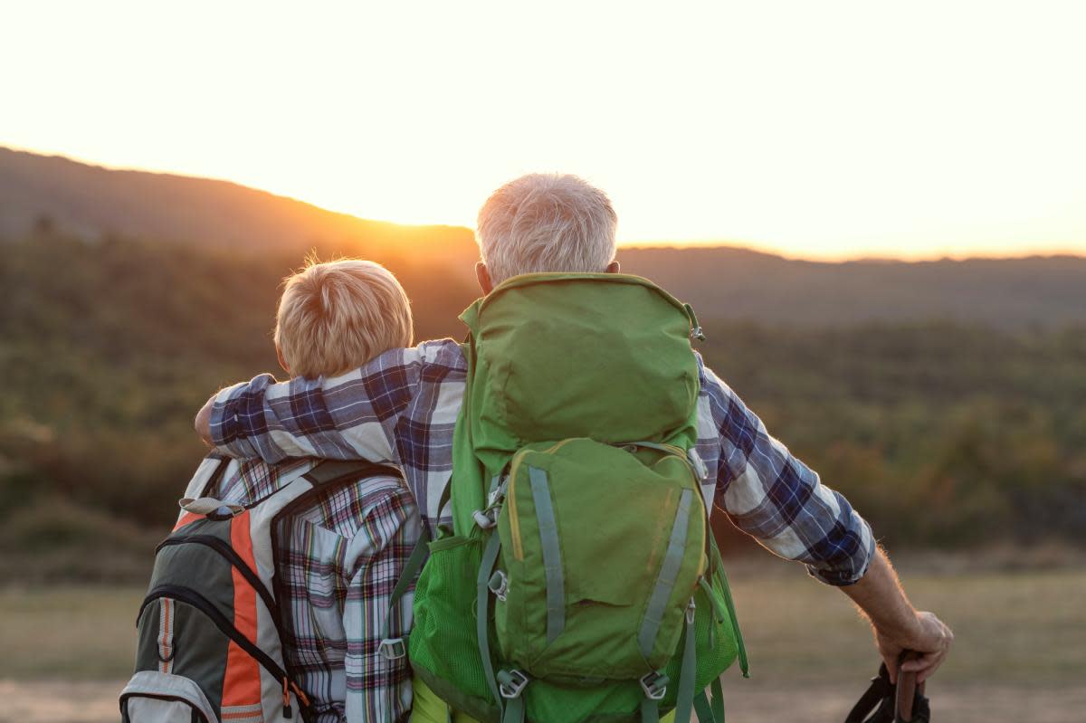 Rear view of two hikers with plaid shirts and backpacks, the older one's arm around the younger. They're gazing at a sunset over distant hills,