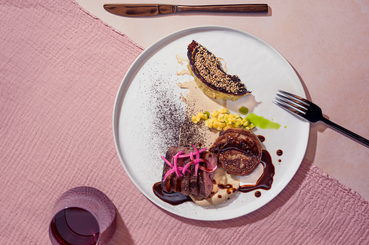 Gourmet dish with sliced meat, pink pickled onions, creamy sauce, roasted vegetables, and sesame-topped wedge on a white plate; wine glass nearby.